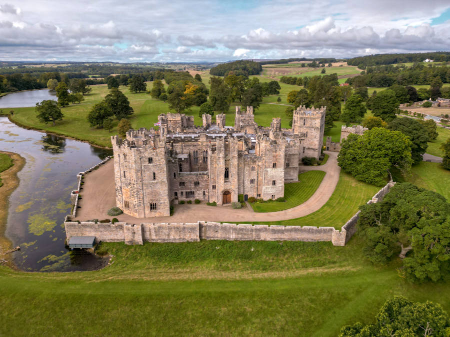 Luftaufnahme von Raby Castle, einer großen steinernen Festung, die von einem Wassergraben, Bäumen und Grasfeldern umgeben ist, unter einem teilweise bewölkten Himmel.