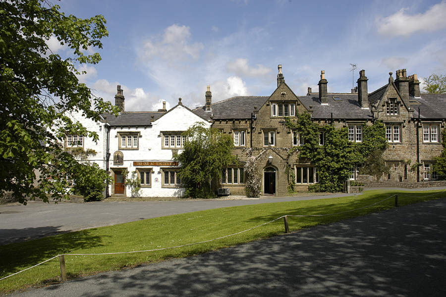 The Inn at Whitewell, a historic stone building with multiple chimneys and ivy-covered walls, stands beside a white-painted structure on a green lawn under a partly cloudy sky.