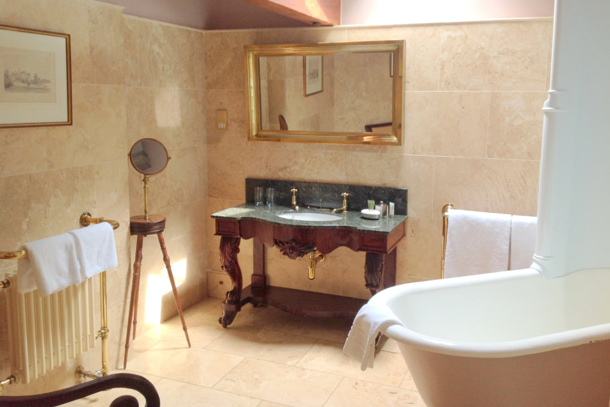 Badezimmer im The Inn at Whitewell mit Steinfliesenwänden, einer freistehenden Badewanne, einem verschnörkelten Holzwaschtisch mit Doppelwaschbecken, einem großen Spiegel und Handtuchhaltern.