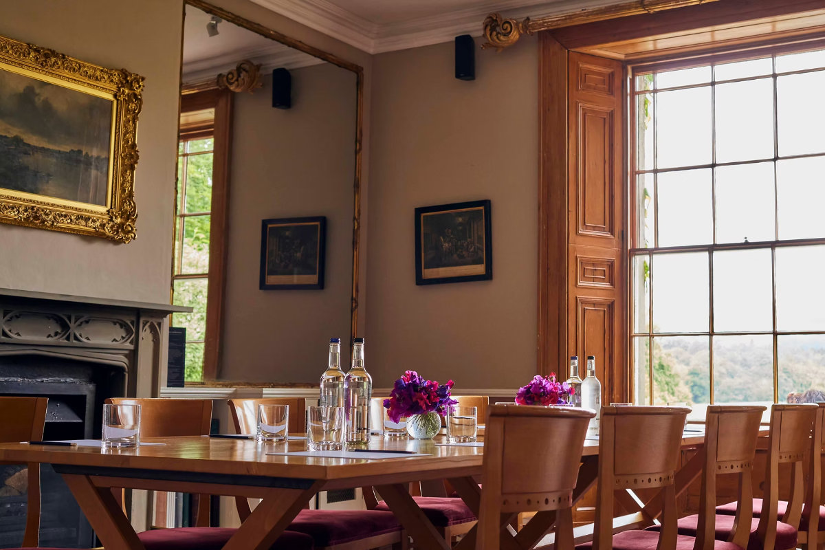 Speisesaal im The Inn at Whitewell mit Holztischen und -stühlen, Glasflaschen, Gläsern, violetten Blumen, einem großen Fenster mit Holzrahmen, Wandspiegeln, Gemälden und einem gemütlichen Kamin.