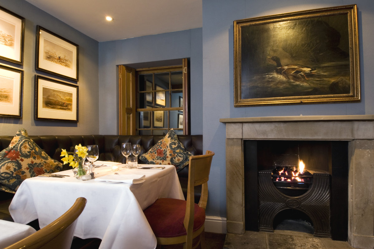 Der gemütliche Restaurantbereich im The Inn at Whitewell verfügt über einen gedeckten Tisch, gepolsterte Sitzgelegenheiten, einen glühenden Kamin und gerahmte Kunstwerke an den eleganten blauen Wänden.