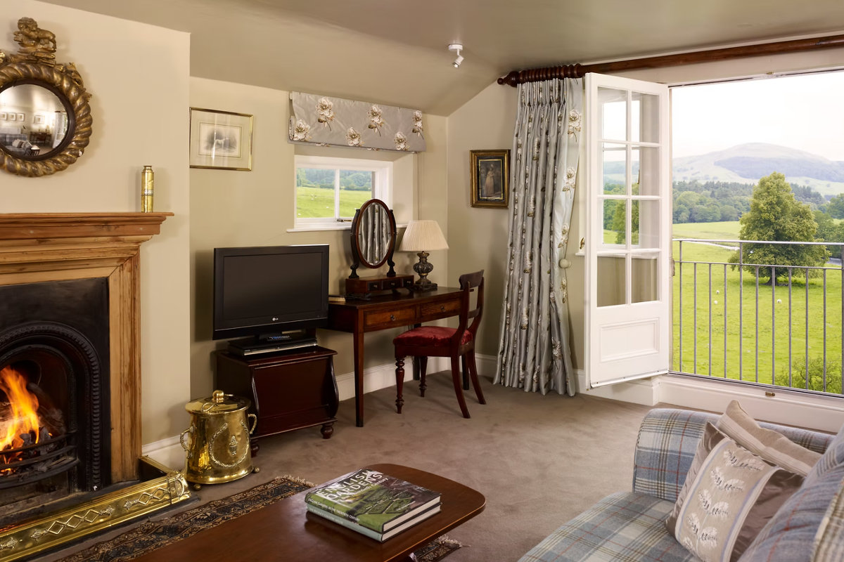 Das gemütliche Wohnzimmer im The Inn at Whitewell verfügt über einen Kamin, einen Fernseher, einen Schreibtisch und offene Flügeltüren, die auf einen Balkon mit Blick auf grüne Felder und ferne Hügel führen.