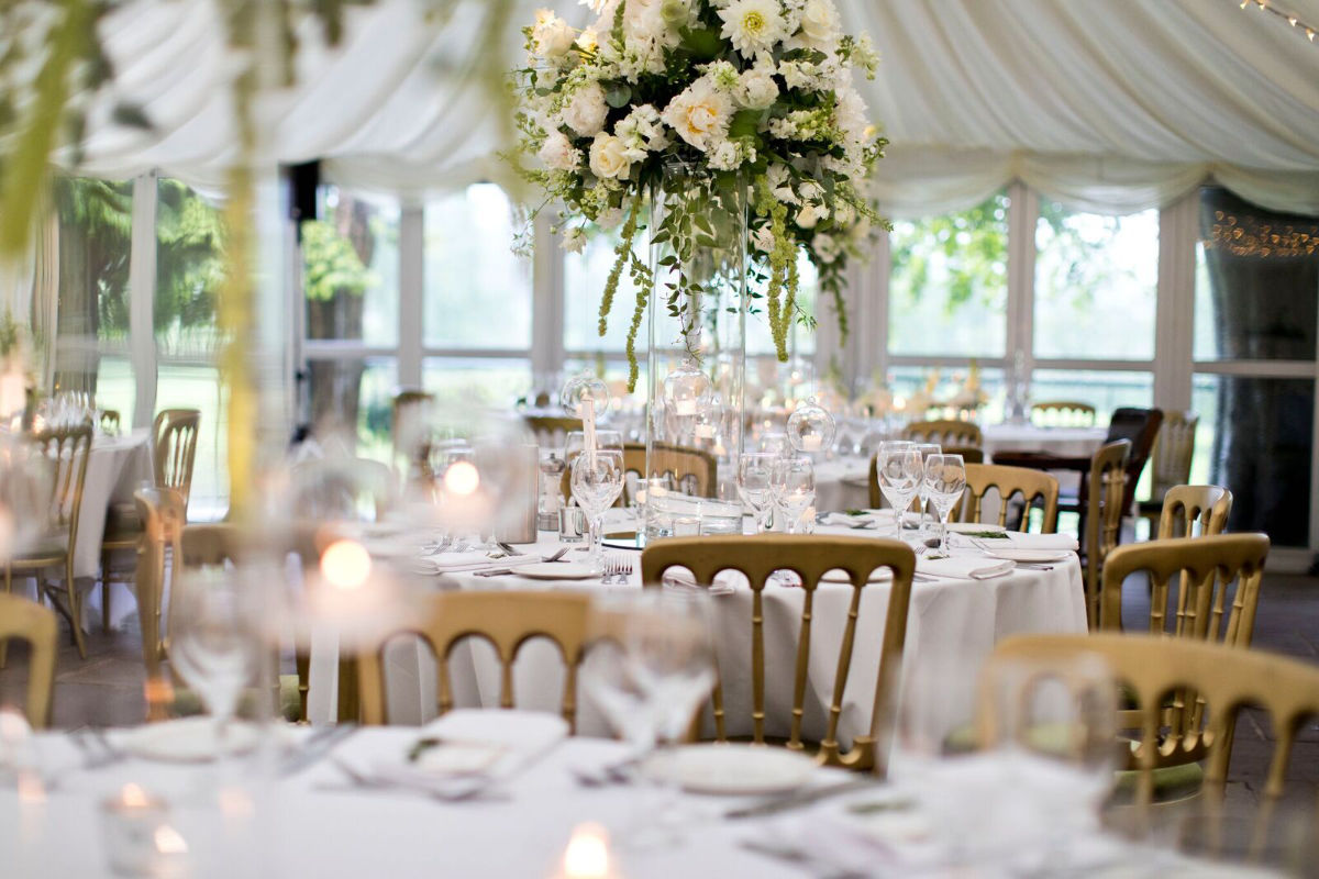 Eleganter Bankettsaal im Inn at Whitewell für eine Veranstaltung mit runden Tischen mit weißen Tischtüchern, goldenen Stühlen und einem hohen Blumenschmuck mit weißen Blumen und Grünzeug.