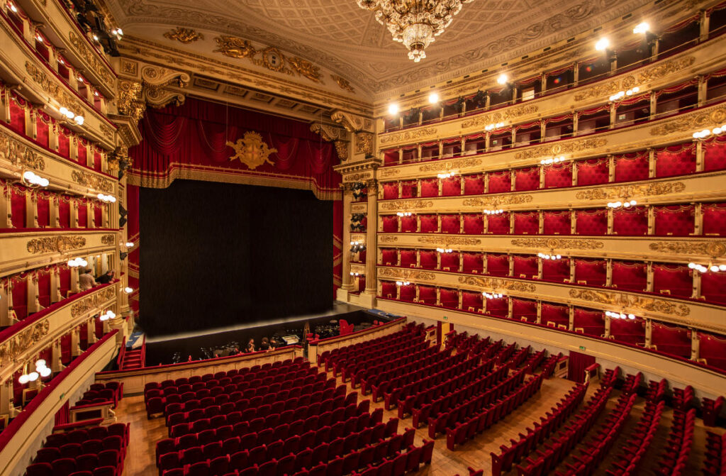 Patrimonio musical europeo: ciudades que cantan 5 La Scala de Milán, opulento interior de teatro con butacas de terciopelo rojo, balcones dorados ornamentados, lámparas de araña y un telón de boca cerrado bajo una cálida iluminación.