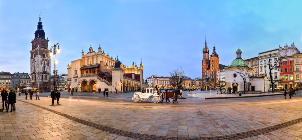 Panoramic view of Krakow’s main square at dusk, featuring St. Mary's Basilica, Cloth Hall, a horse-drawn carriage, and people walking on cobblestone paths.