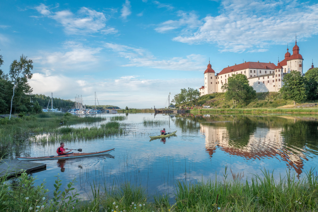 Zwei Personen fahren Kajak auf einem ruhigen See in der Nähe eines Stegs, mit Schloss Läckö und Booten im Hintergrund unter einem blauen Himmel mit vereinzelten Wolken.