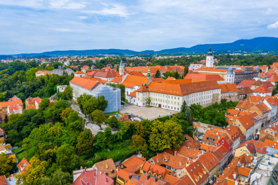 Aerial view of Zagreb Upper Town with red-tiled rooftops, historic buildings, green trees, and distant hills under a partly cloudy sky.