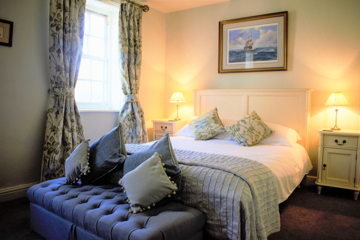 Un letto matrimoniale ben fatto con cuscini floreali, comodini con lampade, una panca imbottita ai piedi del letto e una finestra con tende a fantasia evocano il fascino invitante del White Horse and Griffin.