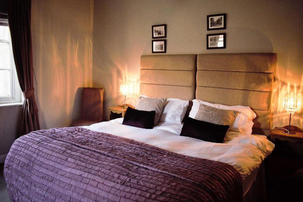 Un letto matrimoniale ben fatto con una testiera imbottita beige alla White Horse e Griffin, due cuscini neri e un copriletto viola; due comodini con lampade e quadri incorniciati alla parete sopra il letto.