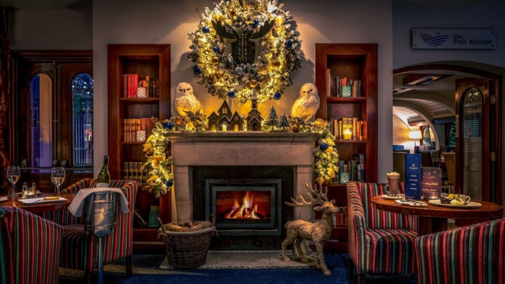 Sapori festivi: cene natalizie negli hotel storici 6 Un ristorante illuminato da luci calde offre una cena natalizia accanto a un caminetto decorato con ghirlande, ornamenti, due figure di gufi bianchi, scaffali e un fuoco scoppiettante. Sedie a strisce e tavoli apparecchiati creano un'accogliente atmosfera festosa in primo piano.