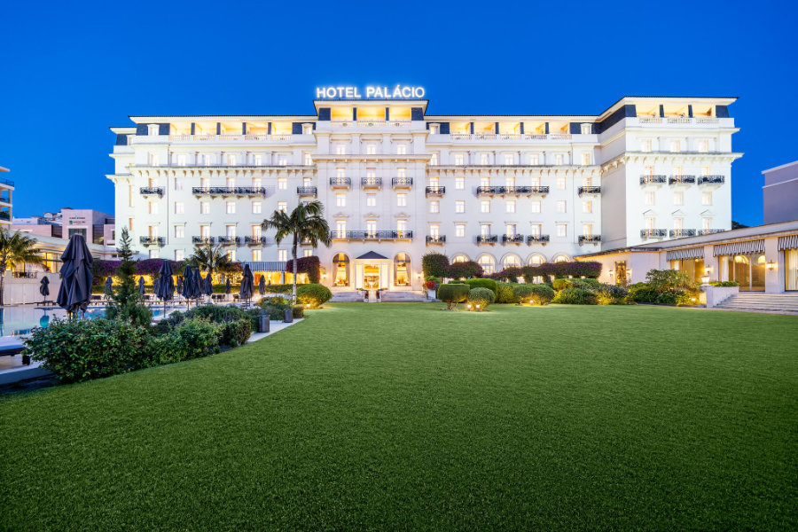 Un hotel grande y bien iluminado llamado "Hotel Palácio Estoril" se alza tras un césped y unos jardines cuidados al atardecer, con mesas al aire libre y sombrillas visibles a la izquierda.