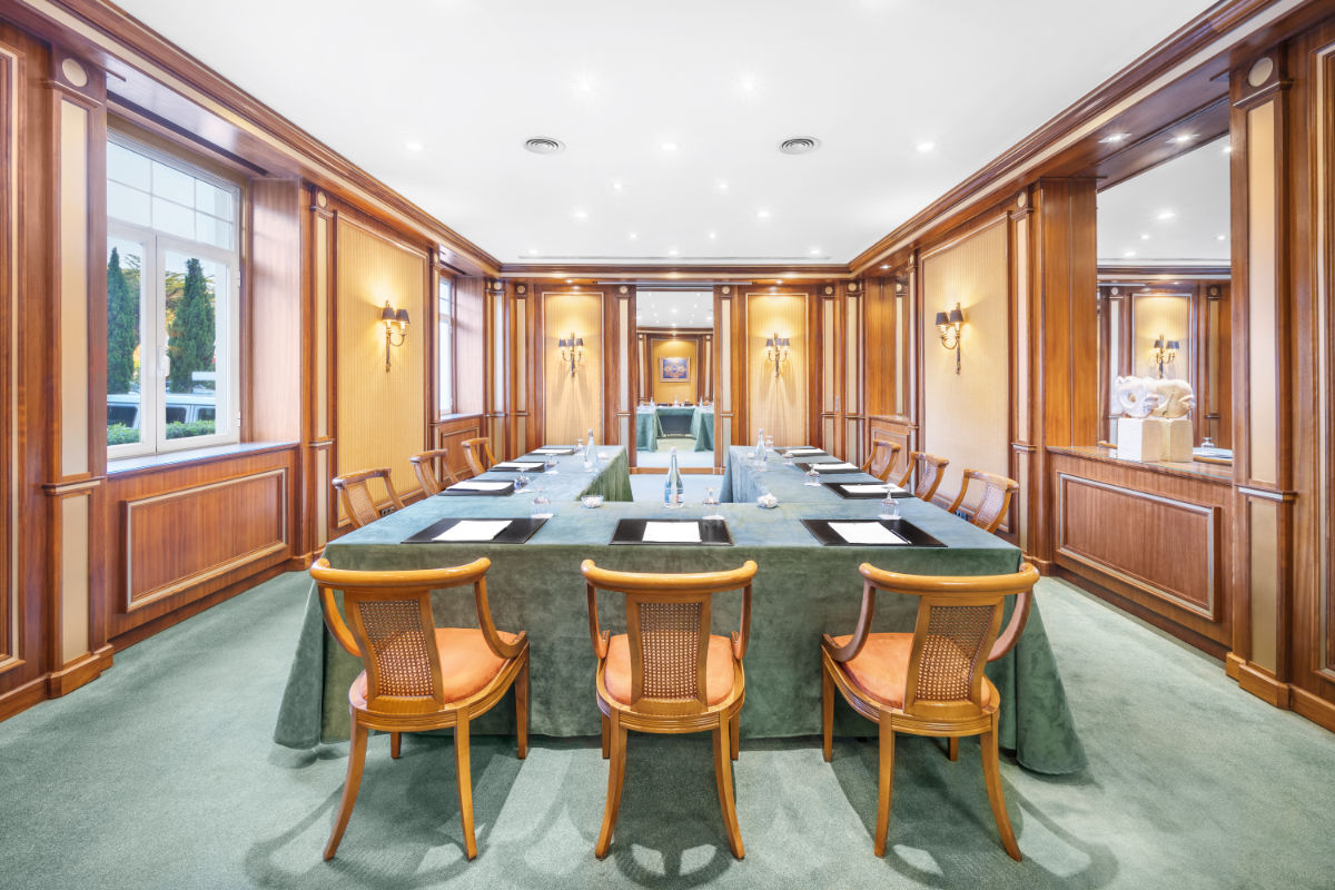 A formal meeting room at Hotel Palácio Estoril features a large square conference table, wooden chairs, green carpet, wood-paneled walls, and natural light streaming through the windows.