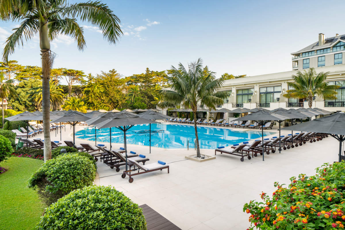 Von Bowie bis Bond: Berühmte Hotels in Europa, die Berühmtheiten lieben 1 Swimmingpool im Freien, umgeben von Liegestühlen und Sonnenschirmen, mit Palmen, Grünflächen und dem großen Hotel Palácio Estoril im Hintergrund unter einem teilweise bewölkten Himmel.