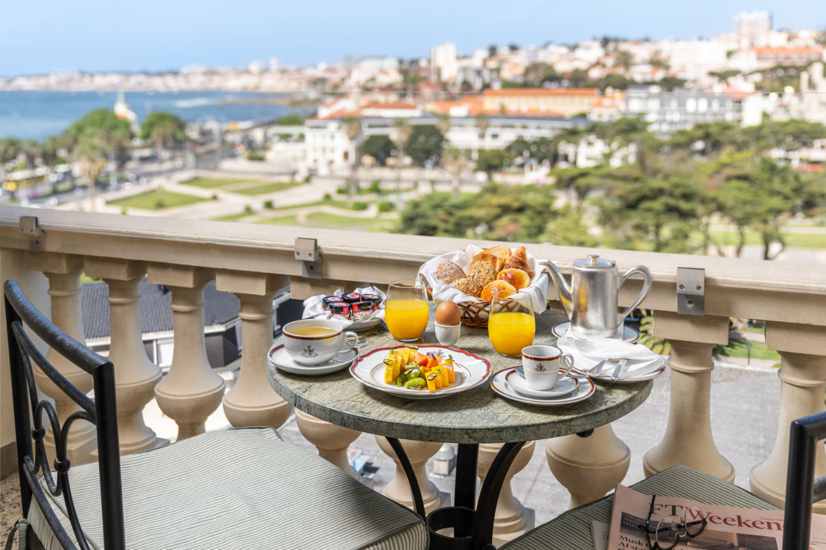 A round table on a balcony at Hotel Palácio Estoril with breakfast items—orange juice, coffee, pastries, and fruit—overlooks a cityscape and waterfront.