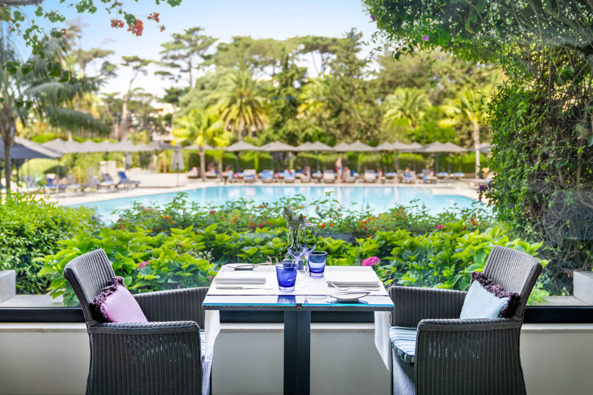 A table set for two with wicker chairs on a terrace at Hotel Palácio Estoril, overlooking a swimming pool and surrounded by lush greenery and trees in the background.