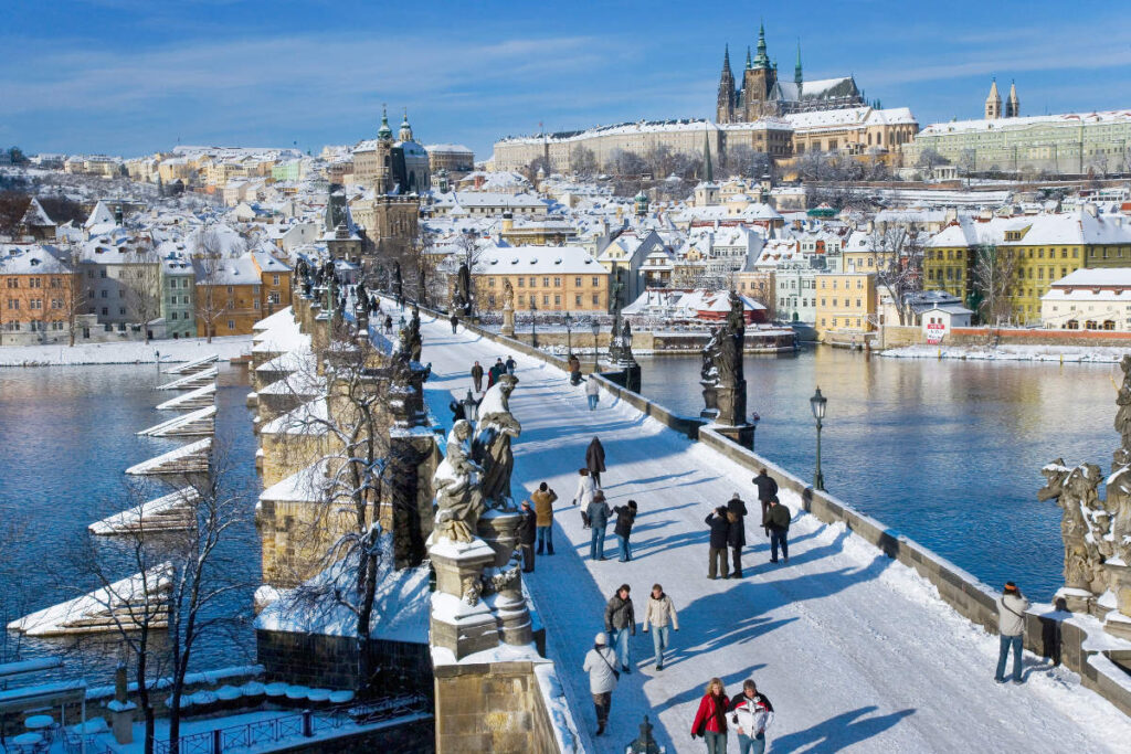 Viajes de invierno en tren: hoteles históricos a lo largo de las rutas ferroviarias europeas 11 La gente camina por el Puente de Carlos, cubierto de nieve, sobre el río Moldava en Praga, con los edificios históricos y el Castillo de Praga al fondo: una escena perfecta para vivir antes o después de los Viajes en tren por Europa en un día despejado de invierno.