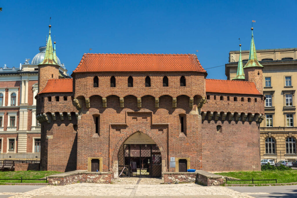 My Town : Kraków - Guide de la ville 1 Une fortification historique en briques avec une entrée en arc, des tours rondes et des flèches à toit vert se dresse fièrement à Cracovie, avec en toile de fond des bâtiments classiques et modernes sous un ciel bleu clair - parfait pour tout guide de Cracovie.