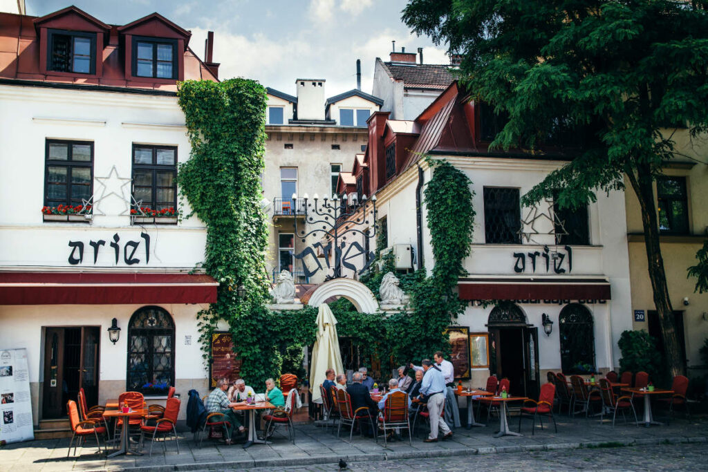 My Town : Kraków - Guide de la ville 4 À Cracovie, des gens s'assoient à des tables en plein air dans un restaurant aux murs blancs, aux chaises rouges et à la façade ornée de lierre ; des panneaux "Ariel" sont affichés et des éléments décoratifs ornent l'entrée - un endroit accueillant figurant dans de nombreux guides de la ville de Cracovie.