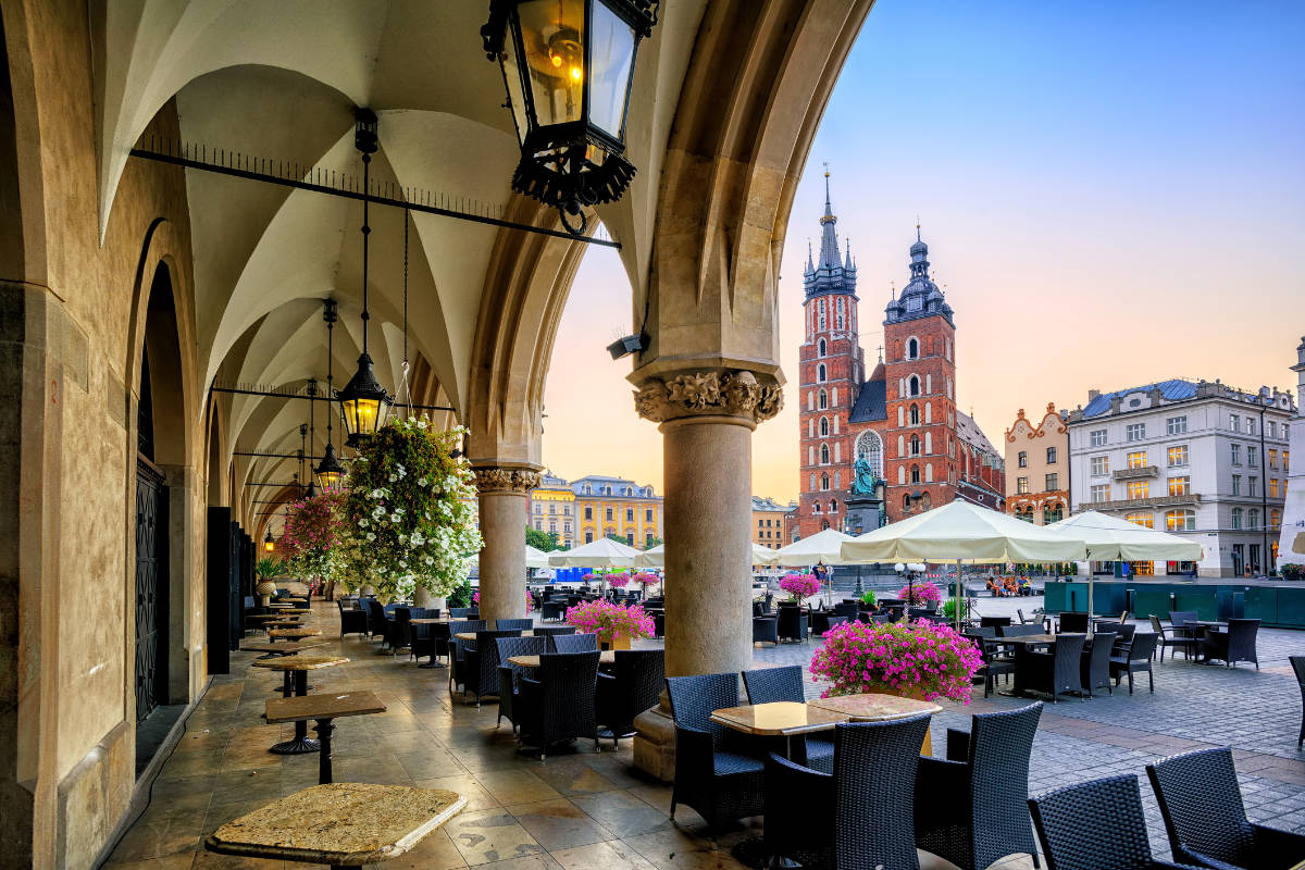 Un caffè all'aperto coperto, con tavoli vuoti e cesti di fiori appesi, si affaccia su una piazza della città di Cracovia, con la Basilica di Santa Maria che risplende al tramonto: una scena perfetta da qualsiasi guida della città di Cracovia.