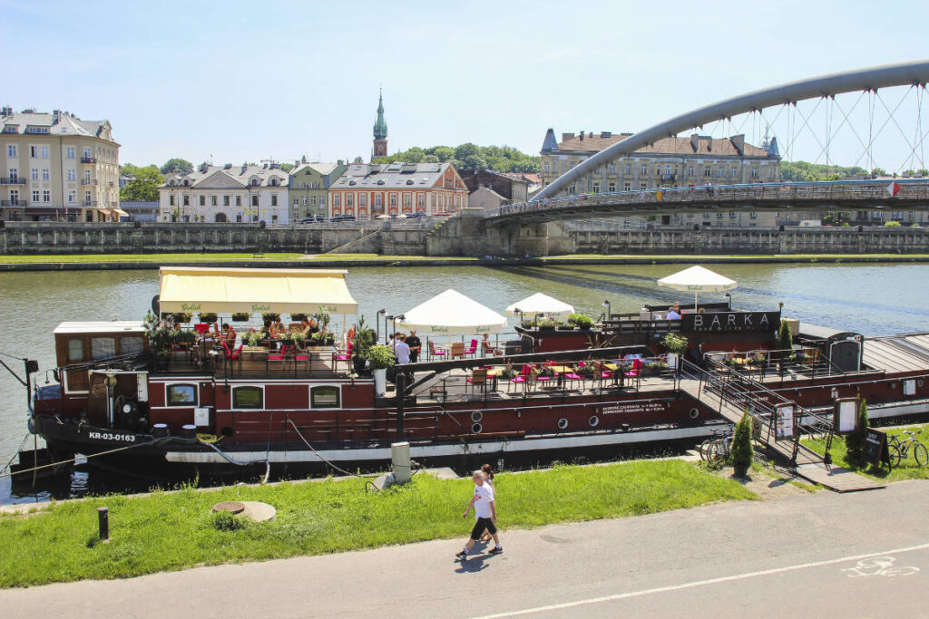 My Town : Kraków - City guide 5 Un bateau-restaurant stationnaire avec des sièges en plein air est amarré au bord de la rivière à Cracovie, avec un piéton marchant le long d'un chemin voisin et un pont en arrière-plan - un endroit idéal figurant dans tous les guides de la ville de Cracovie.