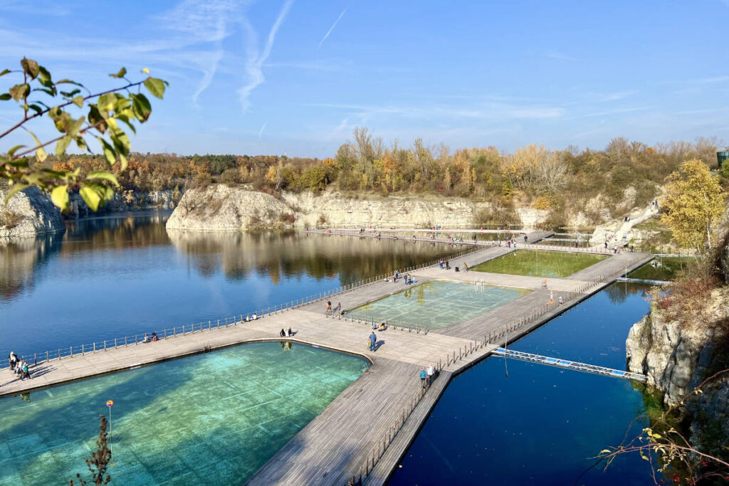 My Town : Kraków - Guide de la ville 8 Une promenade s'étend sur un lac avec des zones de baignade séparées, entouré d'arbres d'automne et de falaises rocheuses sous un ciel bleu - un joyau caché à découvrir dans n'importe quel guide de la ville de Cracovie.