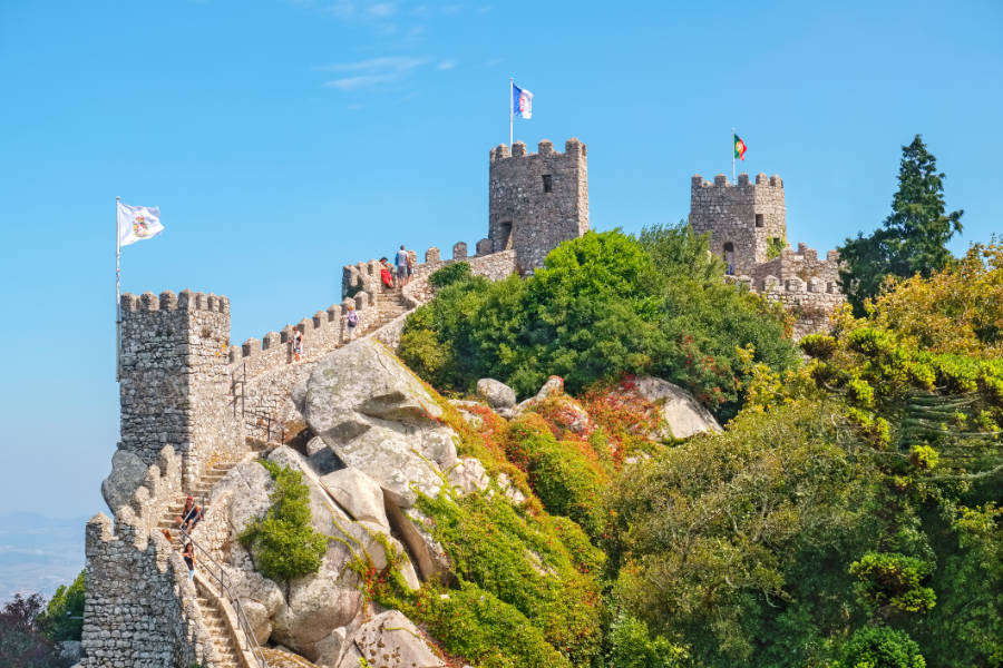 Castello in pietra dei Mori con torri merlate e bandiere in cima a una collina rocciosa, circondato dal verde e visitato dai turisti sotto un cielo azzurro e limpido.