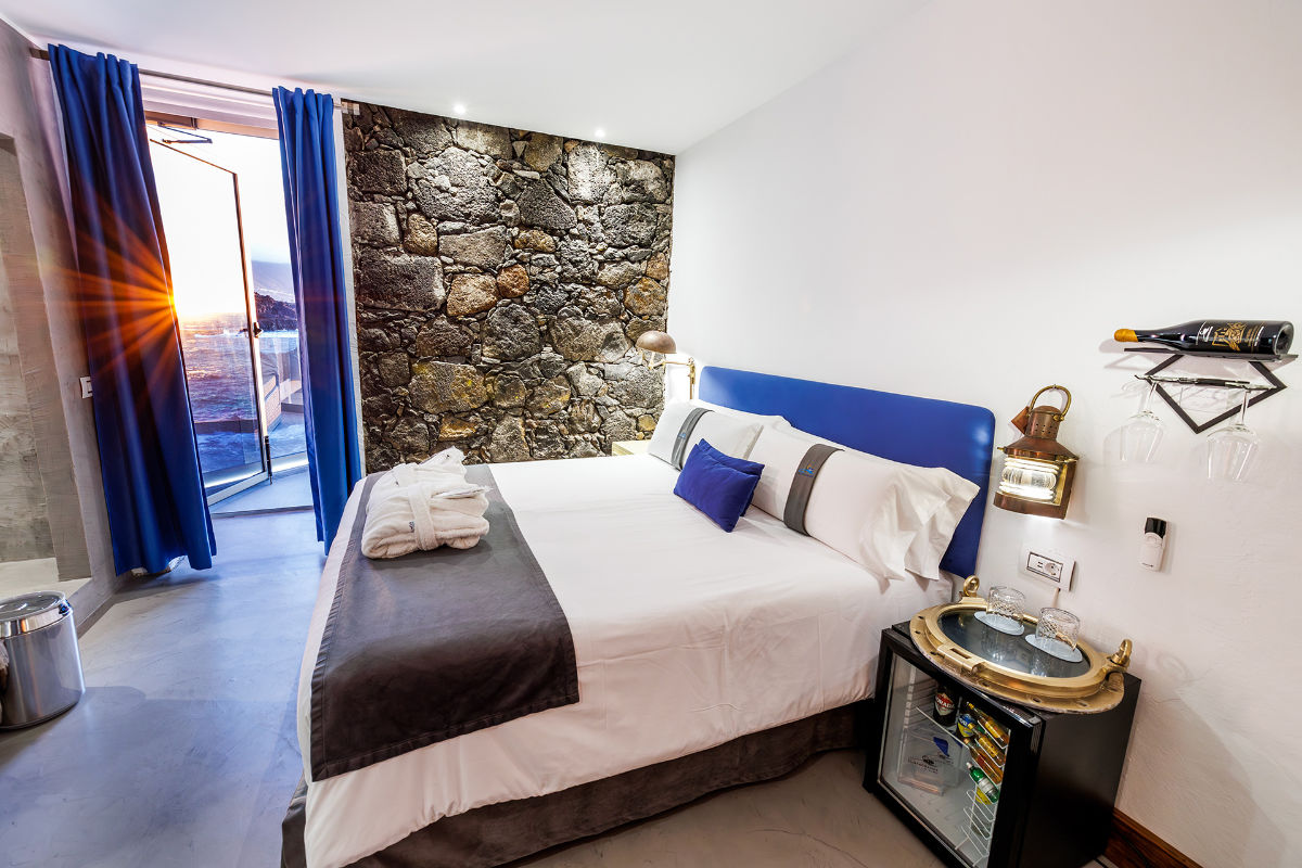 Modern Hotel Puntagrande room with a double bed, stone accent wall, open balcony door for sunset views, minibar, wine shelf, and blue curtains.