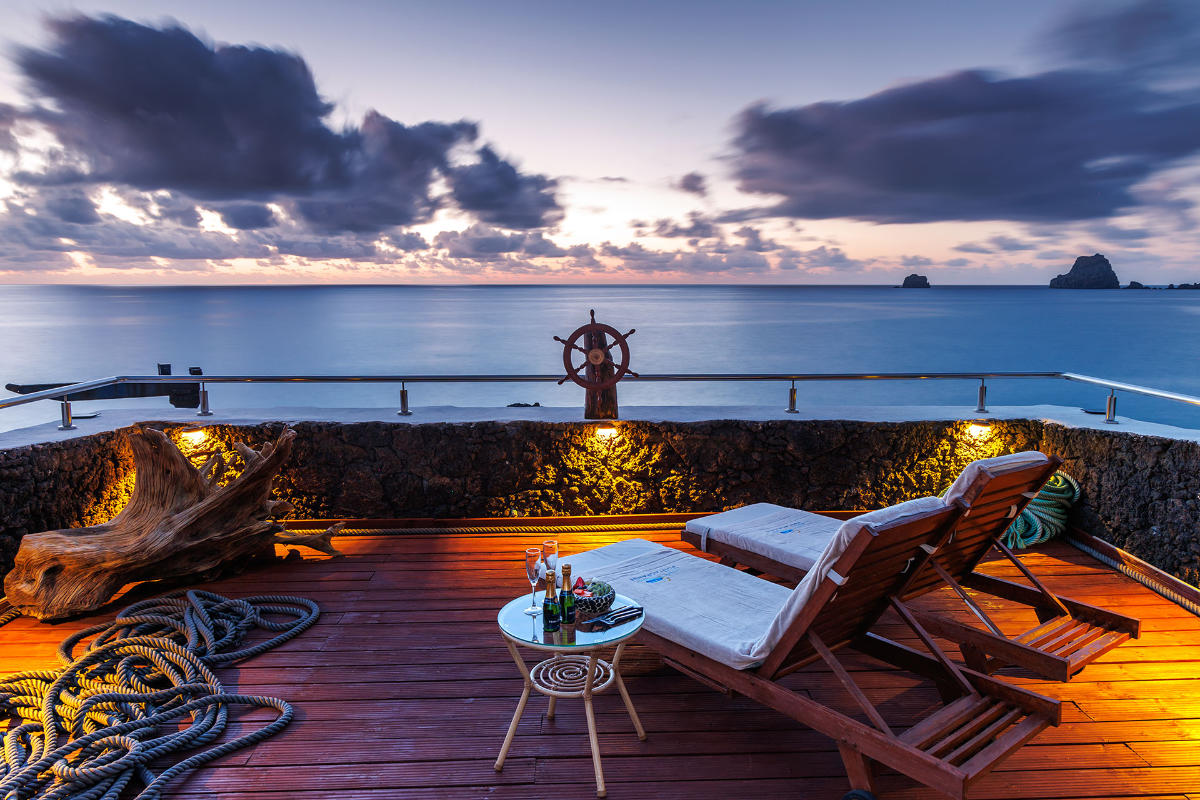 Due sedie a sdraio e un tavolino con bevande su un ponte di legno dell'Hotel Puntagrande si affacciano su un oceano calmo al tramonto, con nuvole drammatiche e rocce lontane all'orizzonte.
