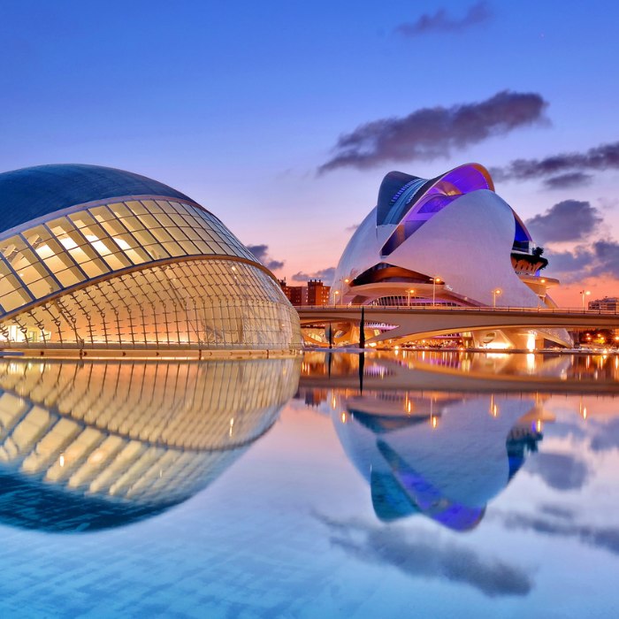 Edificios de arquitectura moderna con cristales curvados y estructuras blancas se reflejan en aguas tranquilas al atardecer bajo un cielo parcialmente nublado.