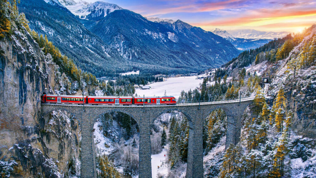 Viajes de invierno en tren: hoteles históricos a lo largo de las rutas ferroviarias europeas 1 Un tren rojo de pasajeros realiza inolvidables viajes en tren mientras cruza un puente viaducto de piedra en un paisaje montañoso nevado al atardecer, rodeado de árboles de hoja perenne y lejanos picos nevados.