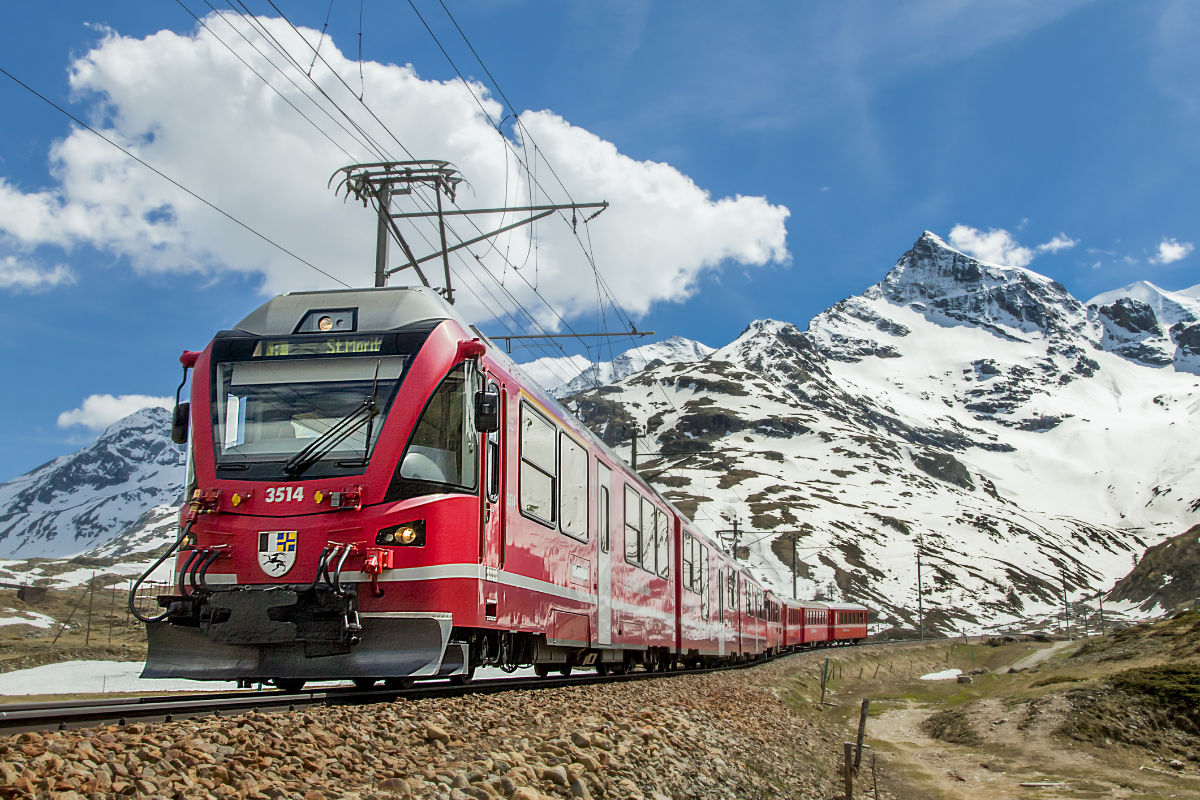 Un train de voyageurs rouge traverse un paysage montagneux avec des sommets enneigés sous un ciel partiellement nuageux, capturant l'esprit des voyages panoramiques en train.
