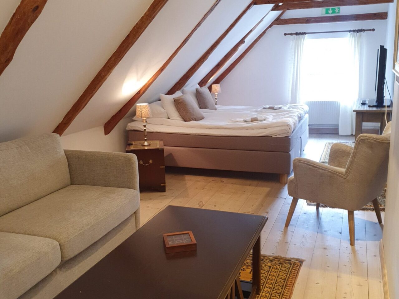 Habitación abuhardillada en Elisefarm con vigas de madera a la vista, cama doble, sofá beige, sillón, mesa auxiliar con lámparas, mesa de centro, TV y ventana con cortinas transparentes que dejan pasar la luz natural.