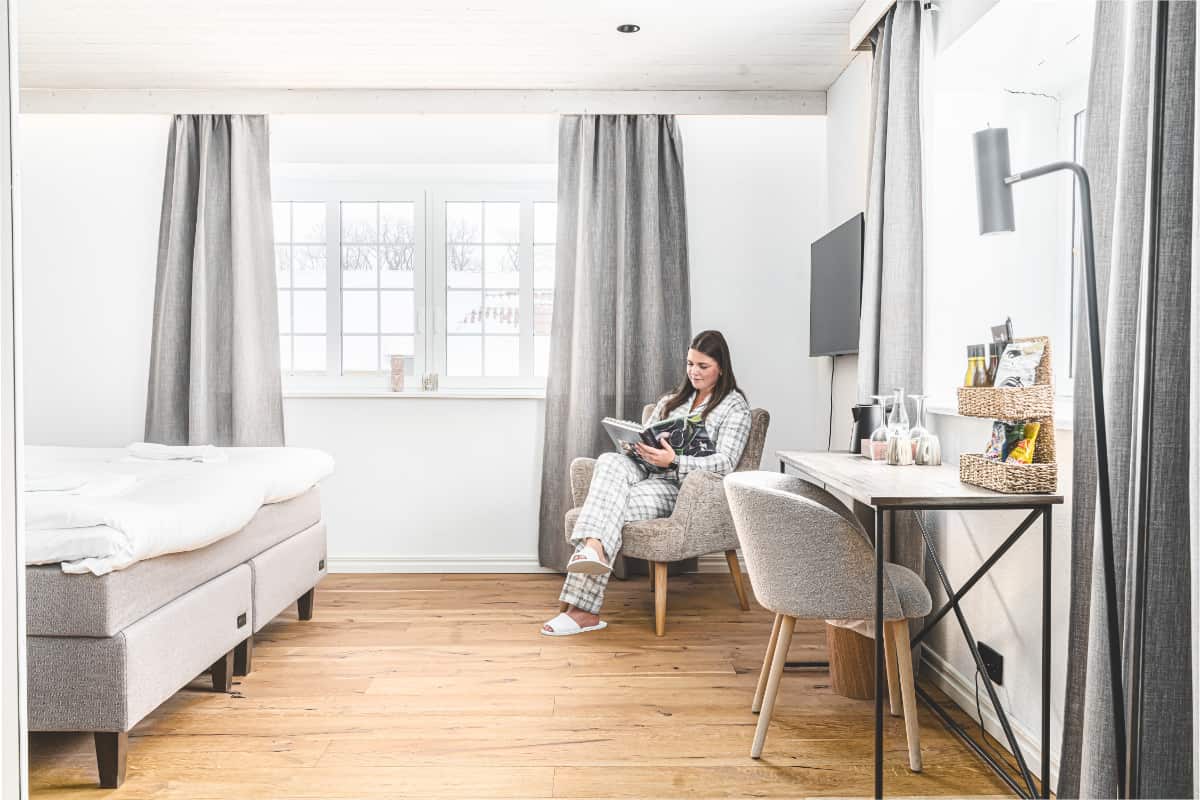 Mujer sentada en una silla junto a una ventana leyendo un libro en un luminoso y moderno dormitorio de Elisefarm con suelo de madera, cama, escritorio y decoración en tonos neutros.