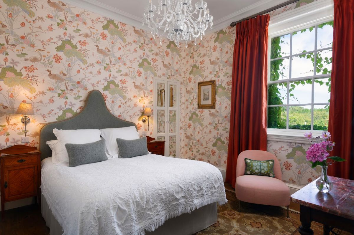La chambre de l'hôtel Dunbrody House comprend un lit double, du papier peint fleuri, deux tables de nuit, un lustre, des rideaux rouges, une fenêtre avec vue sur le jardin, une chaise rose et une petite table avec des fleurs dans ce charmant hôtel 4 étoiles irlandais.