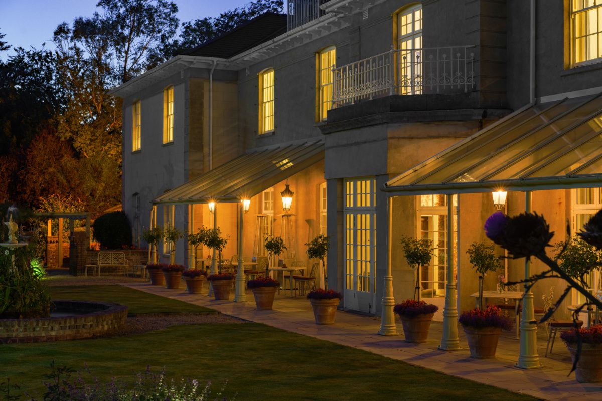 Grande maison aux fenêtres et portes vitrées éclairées, bordée de plantes en pot et de lampes d'extérieur, photographiée au crépuscule avec un jardin paysager au premier plan - parfait pour un séjour à l'hôtel Dunbrody House, un hôtel 4 étoiles de luxe en Irlande.