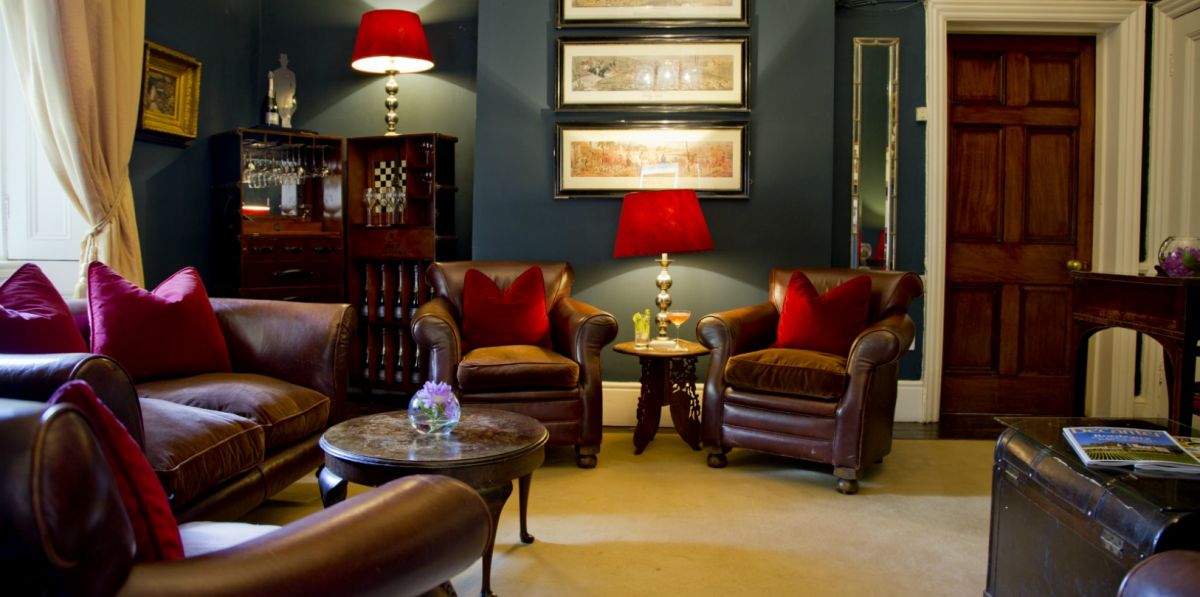 Le salon confortable de l'hôtel Dunbrody House comprend des chaises et un canapé en cuir marron, des coussins rouges, deux lampes rouges, des œuvres d'art encadrées sur des murs sombres et une porte en bois. Il est parfait pour se détendre dans cet élégant hôtel 4 étoiles irlandais.