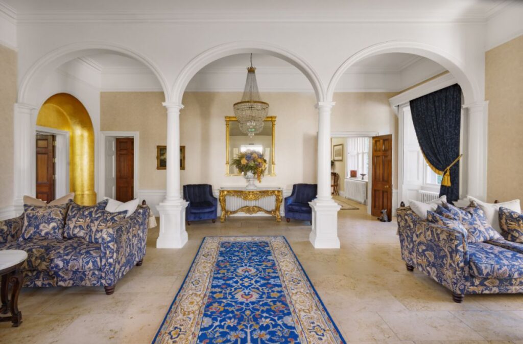 Elegant living room at Dunbrody House Hotel with arched doorways, patterned blue sofas, a blue and gold rug, chandelier, and a gold console table with a flower arrangement below a large mirror—true 4 star hotel Ireland charm.