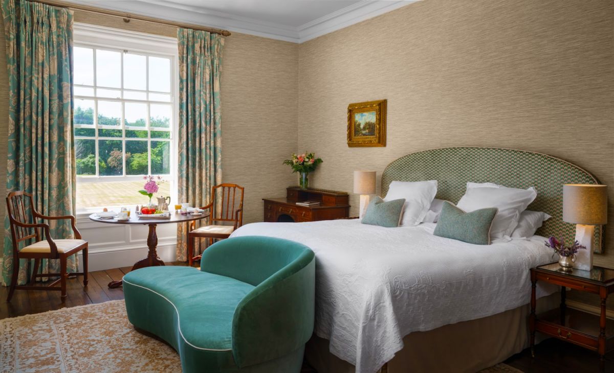 La chambre bien éclairée de l'hôtel Dunbrody House comprend un grand lit, un canapé vert, des meubles en bois et deux chaises près d'une fenêtre. Le petit déjeuner est servi près de la fenêtre donnant sur une pelouse dans ce charmant hôtel 4 étoiles en Irlande.