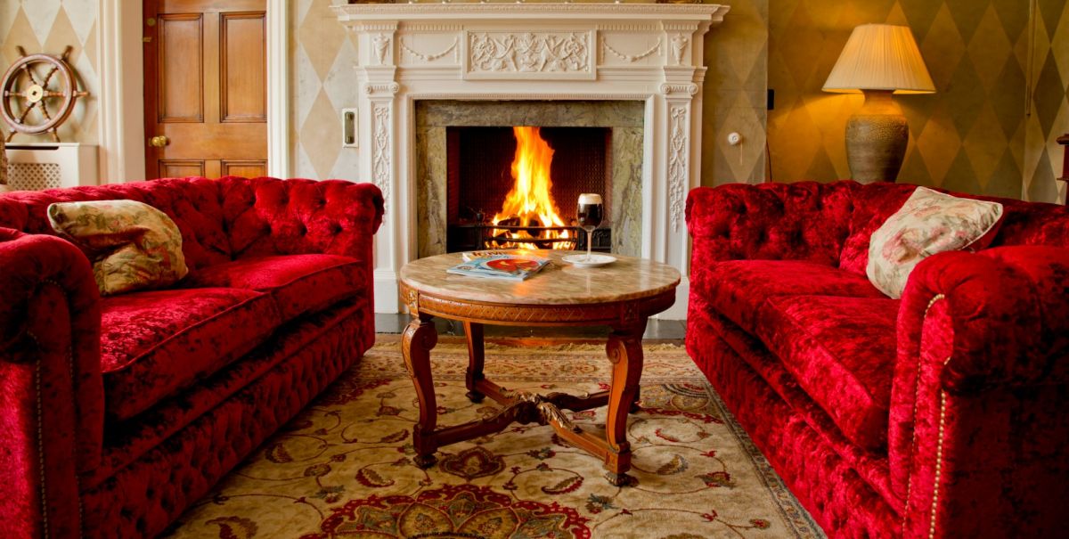 Au Dunbrody House Hotel, deux canapés en velours rouge se font face autour d'une table ronde en bois devant une cheminée allumée ; une lampe et une porte sont visibles à l'arrière-plan de ce charmant hôtel 4 étoiles irlandais.