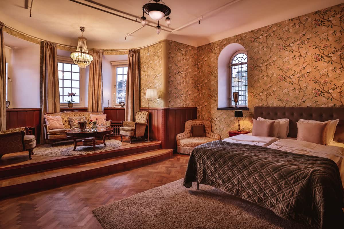 Chambre à coucher à Högberga Gård avec un papier peint à motifs, un lit double, des fenêtres et un coin salon composé de fauteuils, d'un canapé, d'une table basse et d'un lustre sur une plate-forme surélevée. Un éclairage d'ambiance chaleureux renforce l'atmosphère accueillante.