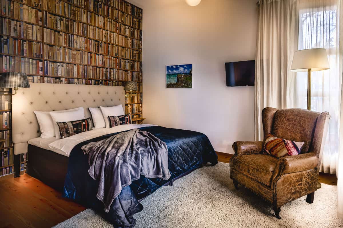 Une chambre confortable à Högberga Gård avec un lit double, un mur tapissé d'une bibliothèque, un fauteuil, une lampe sur pied, une télévision et de grandes fenêtres avec des rideaux transparents.