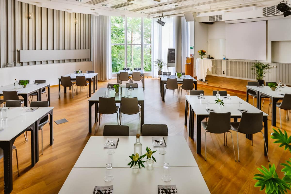 Salle de conférence lumineuse à Högberga Gård avec plusieurs tables et chaises disposées comme dans une salle de classe, décorées de petites plantes, et de grandes fenêtres laissant entrer la lumière naturelle.