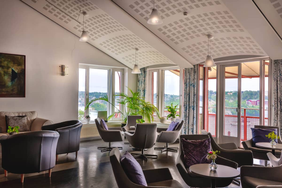 Salon moderne à Högberga Gård avec fauteuils, petites tables, plantes en pot et grandes fenêtres offrant une lumière naturelle et une vue sur les arbres à l'extérieur.