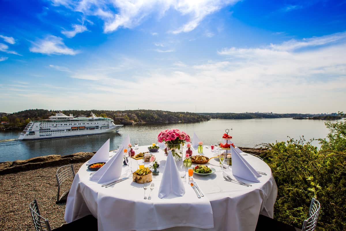 Una mesa redonda al aire libre con comida y bebida en Högberga Gård da a una tranquila masa de agua, con un crucero a lo lejos y un cielo azul en lo alto.
