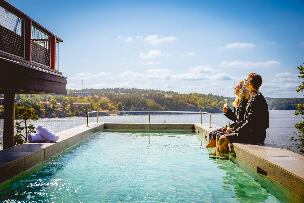 Deux personnes en peignoir sont assises au bord d'une piscine extérieure à Högberga Gård, surplombant un lac, un verre à la main, entourées d'arbres et de ciel bleu.