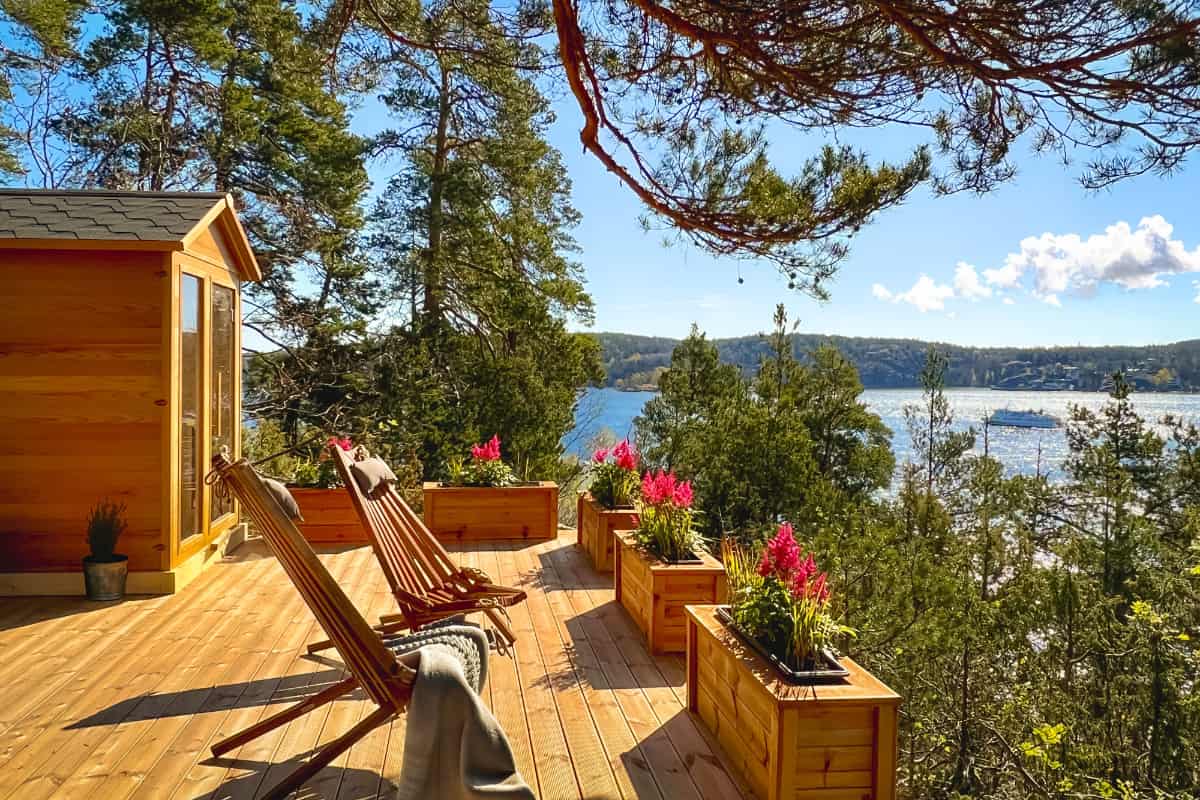 La terrasse en bois de Högberga Gård, équipée de chaises longues et de jardinières, surplombe un lac entouré d'arbres, avec un bateau visible sur l'eau sous un ciel dégagé.