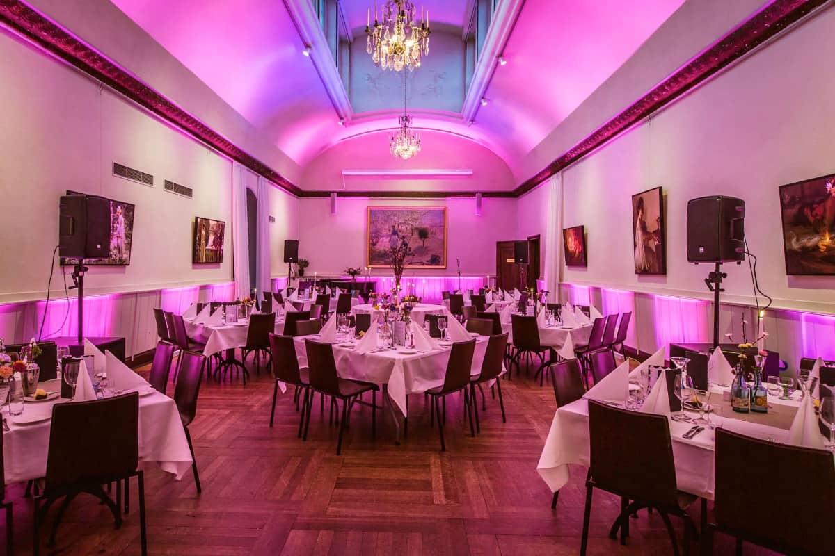 Salle de banquet élégante à Högberga Gård avec un éclairage violet, des tables dressées avec des nappes blanches et des serviettes pliées, un sol en bois, des lustres et des œuvres d'art sur les murs.