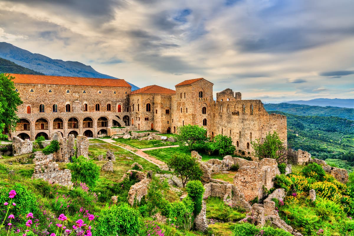 Experiences 1 The large medieval stone fortress of Mystras sits on a hillside, surrounded by green landscape in Greece.