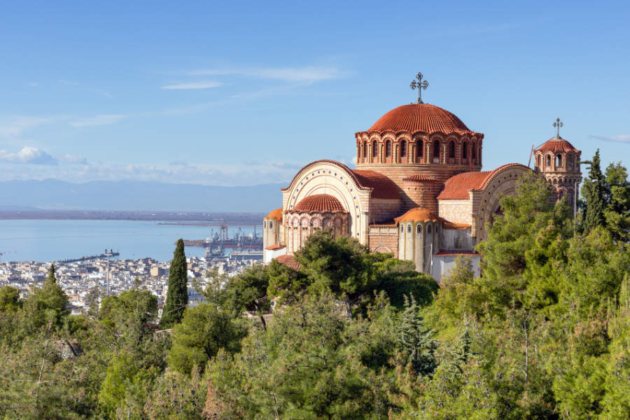 Europe’s Best-Kept Secrets: Lesser-Known Historic Hotel Escapes 5 A large Byzantine-style church with red domes stands on a hill, overlooking a cityscape and harbor by the sea, surrounded by green trees—an enchanting view reminiscent of lesser-known historic hotel escapes.