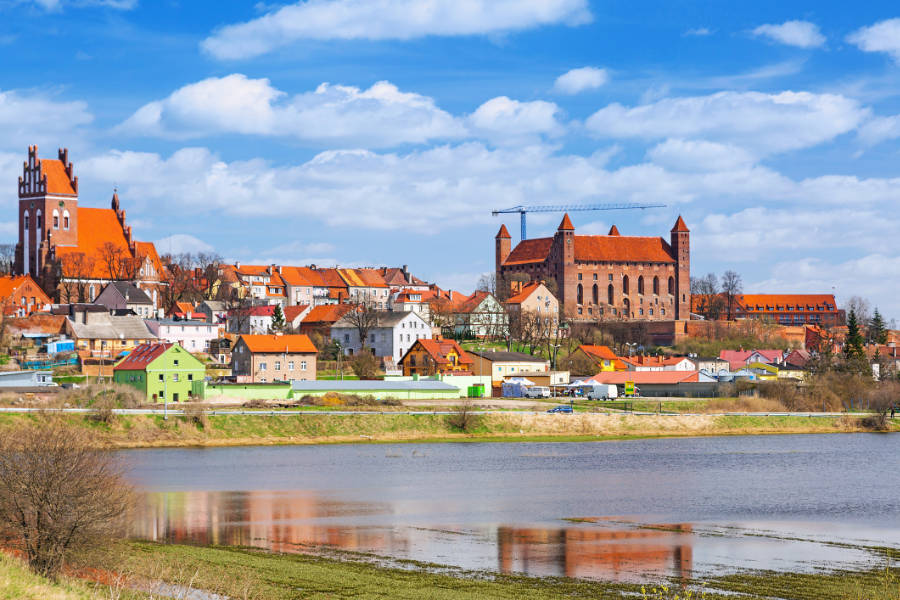 Europe’s Best-Kept Secrets: Lesser-Known Historic Hotel Escapes 19 A town with colorful houses, a large historic castle, and a church sits by a river under a partly cloudy sky—perfect for discovering Lesser-Known Historic Hotel Escapes.